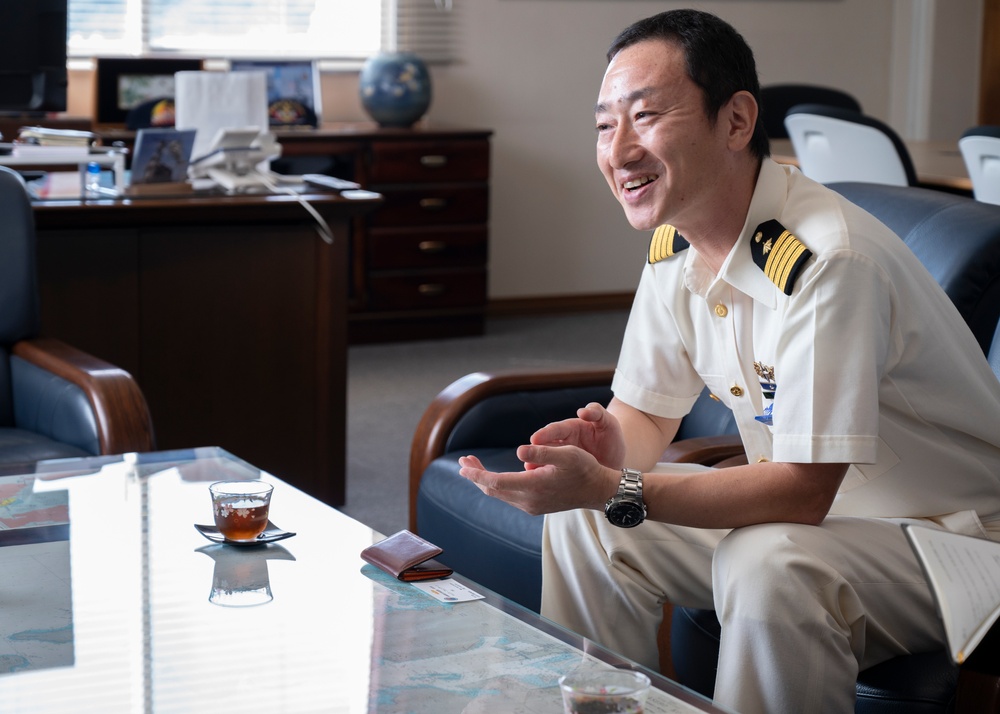 A naval officer in a white dress uniform is seated in what appears to be an office setting, engaged in animated conversation. He is smiling broadly and gesturing with his hands, suggesting a friendly or enthusiastic discussion. The setting includes a glass coffee table with tea and personal items, and a desk with documents and decorative objects in the background.