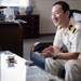 A naval officer in a white dress uniform is seated in what appears to be an office setting, engaged in animated conversation. He is smiling broadly and gesturing with his hands, suggesting a friendly or enthusiastic discussion. The setting includes a glass coffee table with tea and personal items, and a desk with documents and decorative objects in the background.