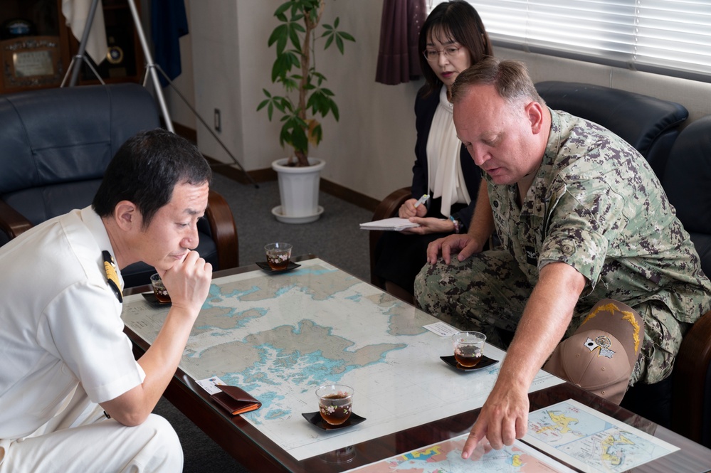 A U.S. Navy servicemember in digital camouflage uniform is pointing at a nautical/geographic map on a table while meeting with a Japanese naval officer in white uniform. A woman, likely serving as an interpreter, stands in the background taking notes. The setting appears to be an official military office, suggesting a bilateral military coordination meeting.