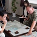 A U.S. Navy servicemember in digital camouflage uniform is pointing at a nautical/geographic map on a table while meeting with a Japanese naval officer in white uniform. A woman, likely serving as an interpreter, stands in the background taking notes. The setting appears to be an official military office, suggesting a bilateral military coordination meeting.