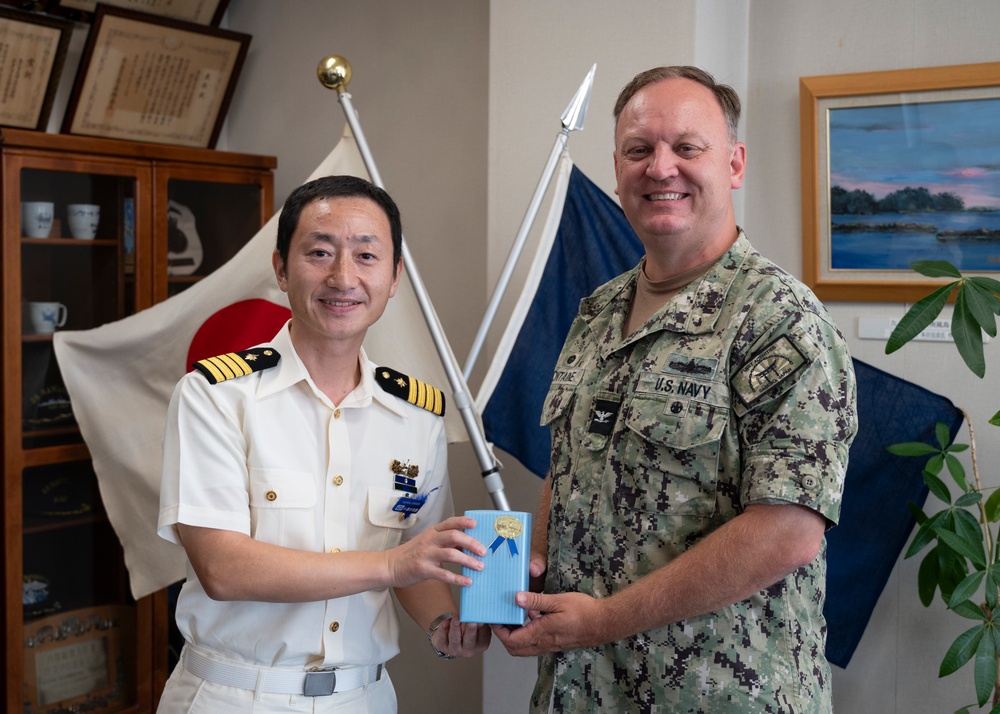 Two military officers, one from the Japan Maritime Self-Defense Force and one from the U.S. Navy, are posing together while jointly holding a gift box with a blue ribbon. The setting appears to be an official office with flags from both Japan and what appears to be a naval command visible in the background. This appears to be a formal bilateral military exchange or courtesy visit.