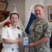 Two military officers, one from the Japan Maritime Self-Defense Force and one from the U.S. Navy, are posing together while jointly holding a gift box with a blue ribbon. The setting appears to be an official office with flags from both Japan and what appears to be a naval command visible in the background. This appears to be a formal bilateral military exchange or courtesy visit.