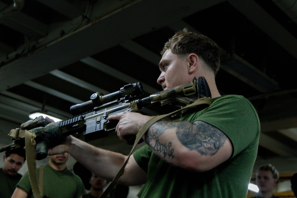 A young male in a green military-style t-shirt is aiming a rifle equipped with an optical scope and sling in what appears to be an indoor training environment. Several other individuals in similar green shirts are visible in the background, suggesting a group training or weapons familiarization session. The setting appears to be inside a ship or military facility based on the industrial ceiling structure.