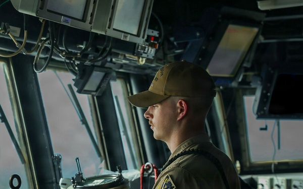 A young male military sailor stands at the helm of a naval vessel's bridge/pilothouse, wearing a tan/khaki uniform with a unit patch on the sleeve and a matching baseball cap. He is looking forward through the bridge windows while operating navigation controls. The setting is clearly a modern warship's command bridge with multiple electronic displays and navigation equipment visible.