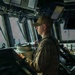 A young male military sailor stands at the helm of a naval vessel's bridge/pilothouse, wearing a tan/khaki uniform with a unit patch on the sleeve and a matching baseball cap. He is looking forward through the bridge windows while operating navigation controls. The setting is clearly a modern warship's command bridge with multiple electronic displays and navigation equipment visible.