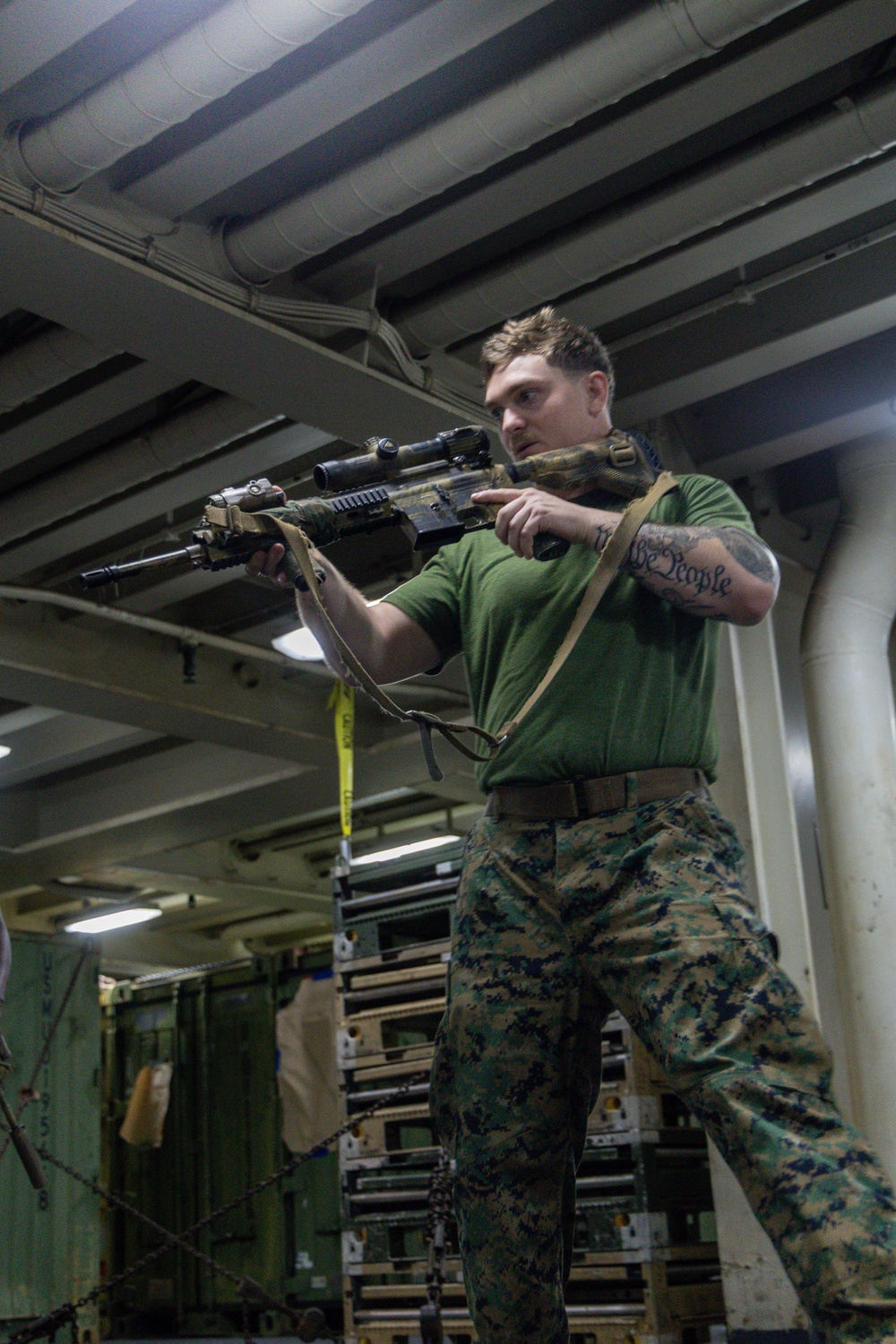 A male service member in U.S. Marine Corps MARPAT digital camouflage trousers and a green t-shirt is holding a rifle with an optic scope and sling in a low-ready or aimed position. The setting appears to be the interior of a naval vessel, identifiable by the overhead pipes, metal bulkheads, and stored military equipment in the background. A tattoo reading 'We People' is visible on his right forearm.