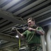 A male service member in U.S. Marine Corps MARPAT digital camouflage trousers and a green t-shirt is holding a rifle with an optic scope and sling in a low-ready or aimed position. The setting appears to be the interior of a naval vessel, identifiable by the overhead pipes, metal bulkheads, and stored military equipment in the background. A tattoo reading 'We People' is visible on his right forearm.
