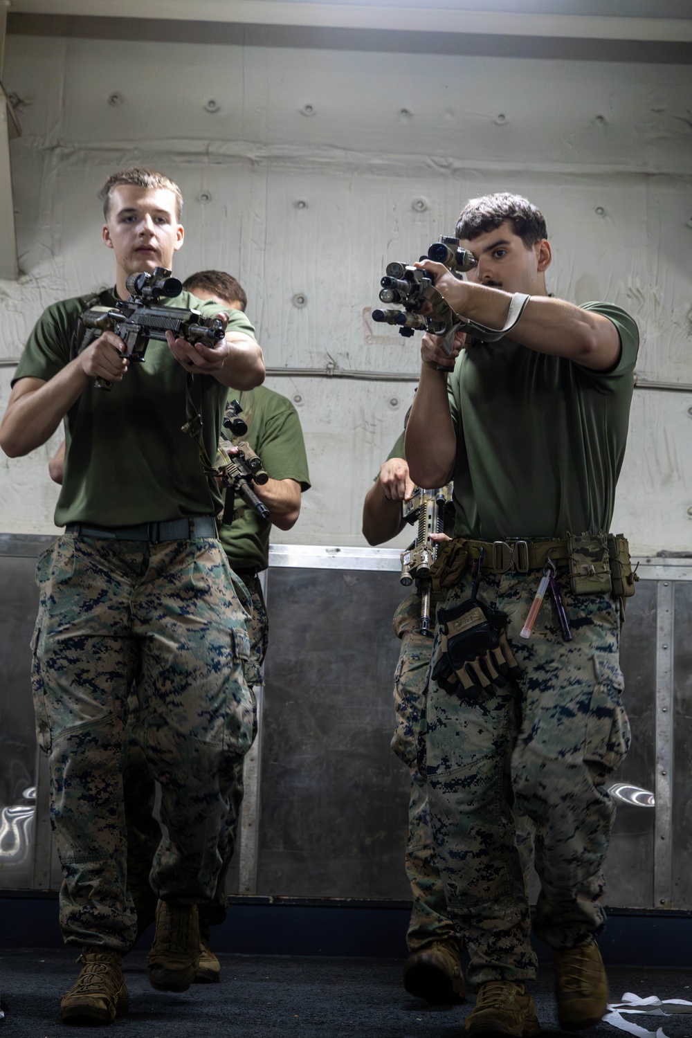 Three military personnel in MARPAT (Marine pattern) camouflage uniforms are conducting what appears to be an indoor close-quarters battle (CQB) training exercise. Two individuals in the foreground are actively aiming rifles equipped with optics and accessories toward the camera, while a third stands in the background. The setting appears to be an indoor training facility or ship compartment.