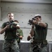 Three military personnel in MARPAT (Marine pattern) camouflage uniforms are conducting what appears to be an indoor close-quarters battle (CQB) training exercise. Two individuals in the foreground are actively aiming rifles equipped with optics and accessories toward the camera, while a third stands in the background. The setting appears to be an indoor training facility or ship compartment.