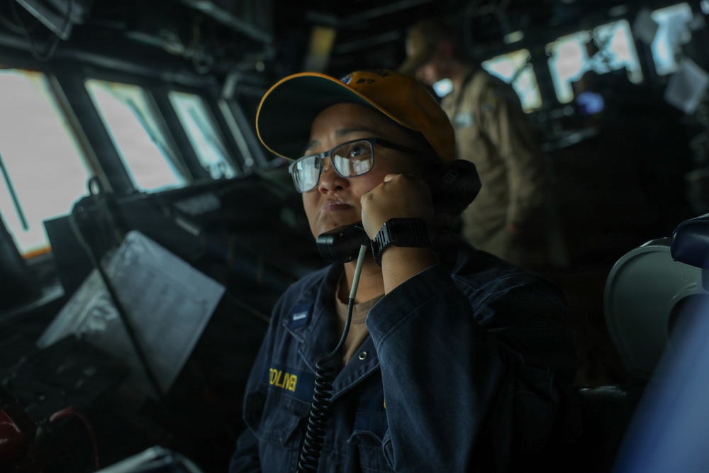 A naval service member is stationed on the bridge of a military vessel, speaking into a sound-powered telephone or handset while monitoring the surroundings. The individual is wearing a dark navy uniform with a name tag partially visible reading 'BOLIN' or similar, along with a yellow/gold baseball cap and glasses. A second crew member in a tan/khaki uniform is visible in the background at what appears to be a navigation or communications station.