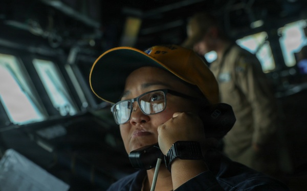 A naval service member is stationed on the bridge of a military vessel, speaking into a sound-powered telephone or handset while monitoring the surroundings. The individual is wearing a dark navy uniform with a name tag partially visible reading 'BOLIN' or similar, along with a yellow/gold baseball cap and glasses. A second crew member in a tan/khaki uniform is visible in the background at what appears to be a navigation or communications station.