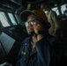 A naval service member is stationed on the bridge of a military vessel, speaking into a sound-powered telephone or handset while monitoring the surroundings. The individual is wearing a dark navy uniform with a name tag partially visible reading 'BOLIN' or similar, along with a yellow/gold baseball cap and glasses. A second crew member in a tan/khaki uniform is visible in the background at what appears to be a navigation or communications station.