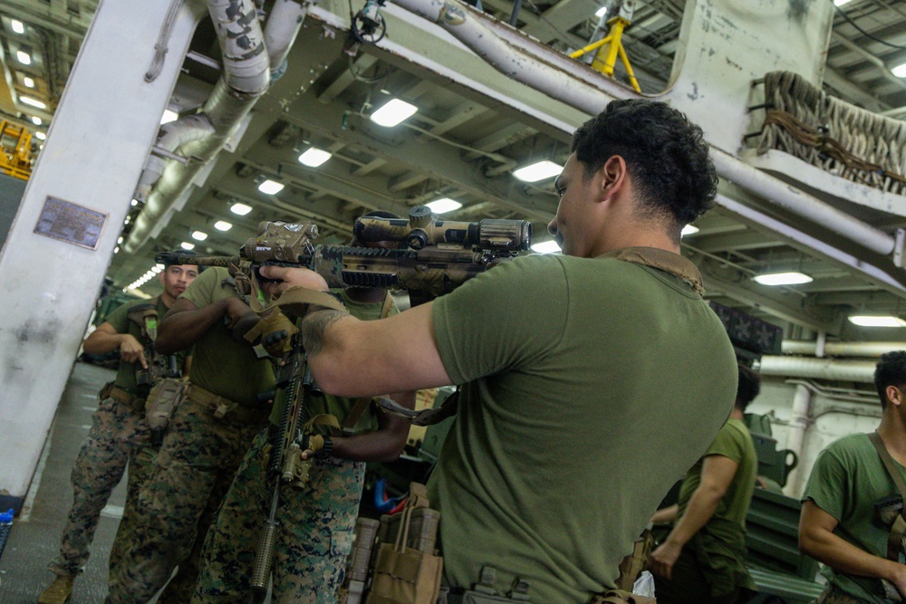 Military personnel, appearing to be U.S. Marines based on their distinctive digital camouflage uniforms, are conducting weapons handling or training exercises inside what appears to be the hangar bay of a naval vessel. The foreground figure in an olive drab t-shirt is aiming a rifle equipped with an optic scope and accessories. Multiple service members are visible in the background observing or participating in the activity.
