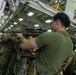 Military personnel, appearing to be U.S. Marines based on their distinctive digital camouflage uniforms, are conducting weapons handling or training exercises inside what appears to be the hangar bay of a naval vessel. The foreground figure in an olive drab t-shirt is aiming a rifle equipped with an optic scope and accessories. Multiple service members are visible in the background observing or participating in the activity.