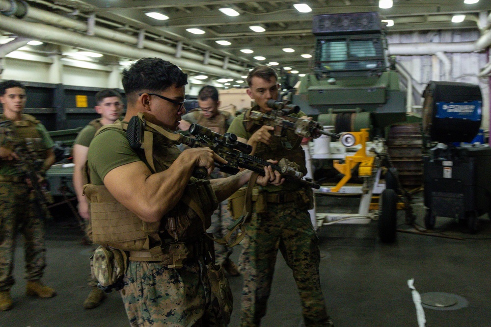Military personnel, appearing to be U.S. Marines based on their MARPAT camouflage uniforms, are conducting weapons handling or training inside what appears to be a ship's well deck or vehicle bay. Two foreground personnel are actively handling scoped rifles while others observe in the background. Military vehicles and equipment are visible in the bay behind them.