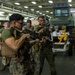 Military personnel, appearing to be U.S. Marines based on their MARPAT camouflage uniforms, are conducting weapons handling or training inside what appears to be a ship's well deck or vehicle bay. Two foreground personnel are actively handling scoped rifles while others observe in the background. Military vehicles and equipment are visible in the bay behind them.
