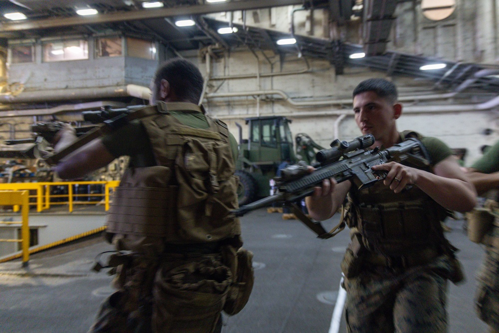 Two military personnel in combat gear and tactical vests are moving through what appears to be a large ship's hangar bay or vehicle deck. The foreground figure is holding an assault rifle with an optical scope in a ready position. The setting contains heavy equipment and industrial infrastructure consistent with a naval vessel's interior.