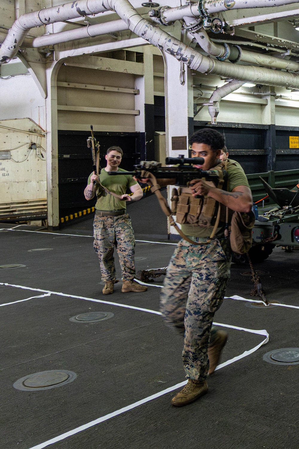 Two U.S. Marines in MARPAT camouflage uniforms are conducting a tactical training drill inside what appears to be the well deck or vehicle bay of a naval amphibious ship. The foreground Marine is in a shooting stance aiming a rifle equipped with a scope and tactical accessories, while the background Marine holds a rifle upright and observes. Military vehicles and ship infrastructure including overhead pipes and tie-down deck fittings are visible in the background.