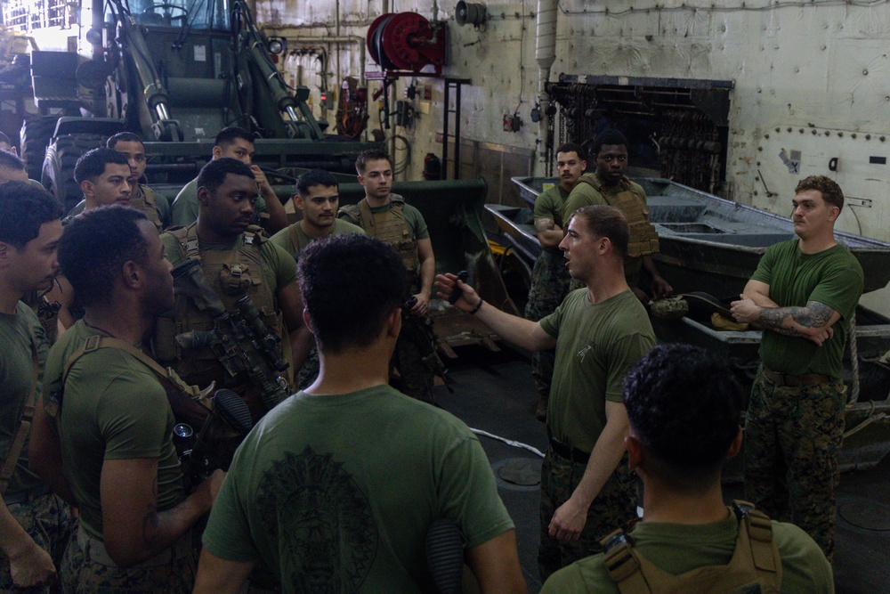 A group of military personnel, appearing to be U.S. Marines based on their olive drab uniforms and tactical gear, are gathered in what appears to be a ship's well deck or vehicle bay. One individual in the center appears to be conducting a briefing or instruction session, gesturing with his hand toward the group. Several flat-bottomed aluminum boats are visible in the background, suggesting preparation for a waterborne operation.