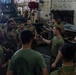 A group of military personnel, appearing to be U.S. Marines based on their olive drab uniforms and tactical gear, are gathered in what appears to be a ship's well deck or vehicle bay. One individual in the center appears to be conducting a briefing or instruction session, gesturing with his hand toward the group. Several flat-bottomed aluminum boats are visible in the background, suggesting preparation for a waterborne operation.