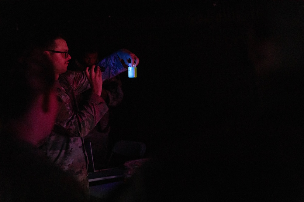 A person wearing camouflage military uniform and glasses is holding up a small glowing container or vial in a very dark environment. The object emits a blue and yellow/green light, suggesting it may be a chemlight, test tube, or similar luminescent item. A second person in camouflage is partially visible in the background.