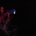 A person wearing camouflage military uniform and glasses is holding up a small glowing container or vial in a very dark environment. The object emits a blue and yellow/green light, suggesting it may be a chemlight, test tube, or similar luminescent item. A second person in camouflage is partially visible in the background.