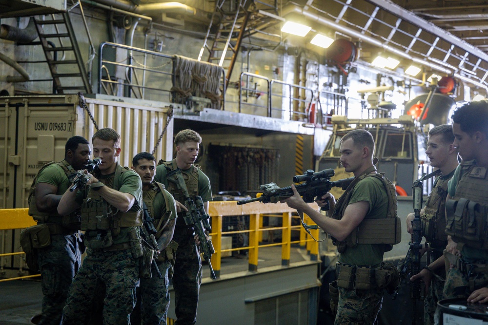 A group of military personnel, appearing to be U.S. Marines based on their camouflage uniforms, are gathered inside what appears to be the well deck of a naval amphibious ship. One Marine is demonstrating or inspecting a scoped rifle while others observe, suggesting a weapons familiarization or training session. A shipping container marked 'USNU 019683' and military vehicles are visible in the background.