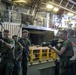 A group of military personnel, appearing to be U.S. Marines based on their camouflage uniforms, are gathered inside what appears to be the well deck of a naval amphibious ship. One Marine is demonstrating or inspecting a scoped rifle while others observe, suggesting a weapons familiarization or training session. A shipping container marked 'USNU 019683' and military vehicles are visible in the background.