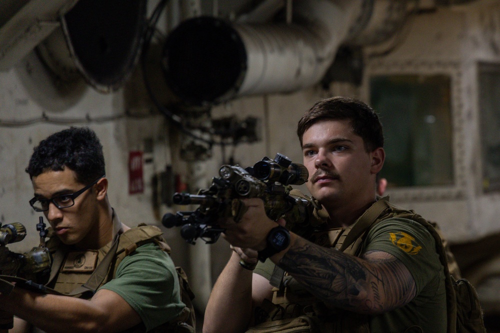 Two military personnel in tactical gear are shown in what appears to be an interior shipboard environment. The foreground individual is aiming a rifle equipped with optical sights and accessories, while the second individual holds a weapon at a lower ready position. Both wear olive drab t-shirts with tan/coyote plate carriers.