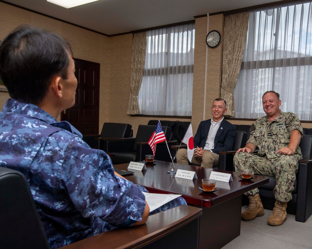 A formal bilateral meeting is taking place between Japanese and U.S. military/civilian representatives in an official conference room. Small American and Japanese flags are displayed on the table, indicating an official diplomatic or military liaison meeting. The participants appear relaxed and engaged in friendly conversation.