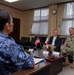 A formal bilateral meeting is taking place between Japanese and U.S. military/civilian representatives in an official conference room. Small American and Japanese flags are displayed on the table, indicating an official diplomatic or military liaison meeting. The participants appear relaxed and engaged in friendly conversation.