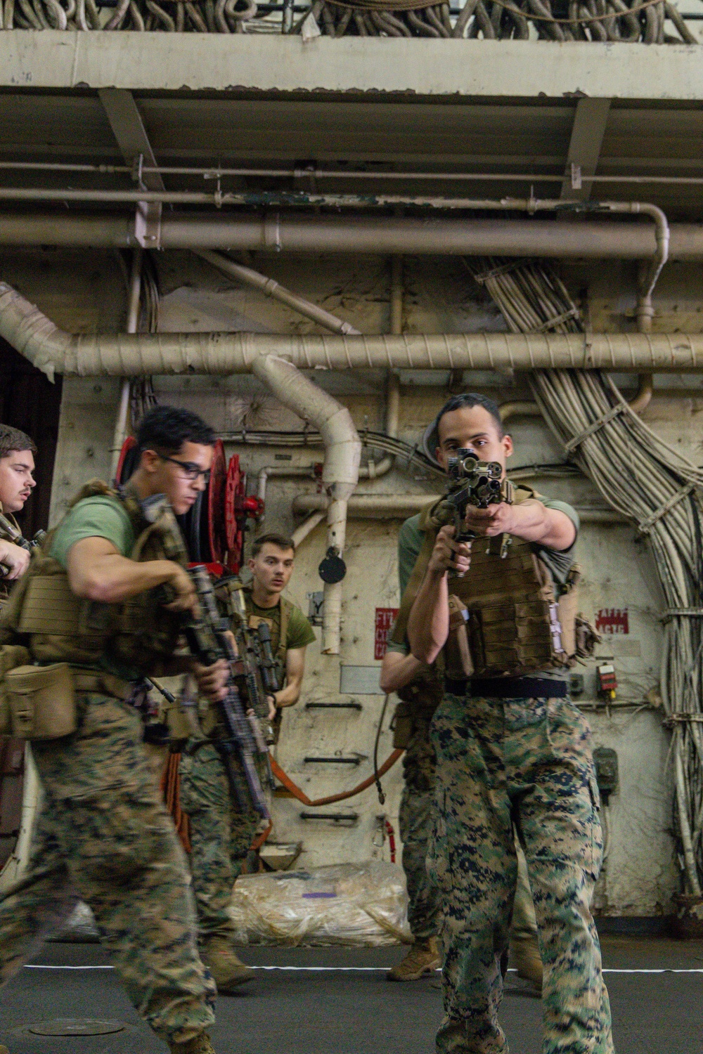 Military personnel, appearing to be U.S. Marines based on their MARPAT camouflage uniforms and plate carrier vests, are conducting a tactical training drill inside what appears to be the hangar bay of a naval vessel. At least four individuals are visible, with two in the foreground actively moving with rifles in ready positions. The industrial interior environment features exposed pipes, cable bundles, and fire hoses consistent with a ship's interior.