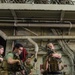 Military personnel, appearing to be U.S. Marines based on their MARPAT camouflage uniforms and plate carrier vests, are conducting a tactical training drill inside what appears to be the hangar bay of a naval vessel. At least four individuals are visible, with two in the foreground actively moving with rifles in ready positions. The industrial interior environment features exposed pipes, cable bundles, and fire hoses consistent with a ship's interior.