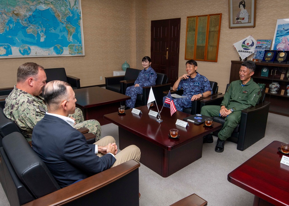 A formal military meeting is taking place in an office setting between what appear to be U.S. Navy personnel and Japanese Self-Defense Force members. Small desktop flags representing Japan and the United States are prominently displayed on the conference table, indicating a bilateral military liaison or coordination meeting. The atmosphere appears relaxed and collegial, with participants engaged in conversation.