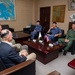 A formal military meeting is taking place in an office setting between what appear to be U.S. Navy personnel and Japanese Self-Defense Force members. Small desktop flags representing Japan and the United States are prominently displayed on the conference table, indicating a bilateral military liaison or coordination meeting. The atmosphere appears relaxed and collegial, with participants engaged in conversation.