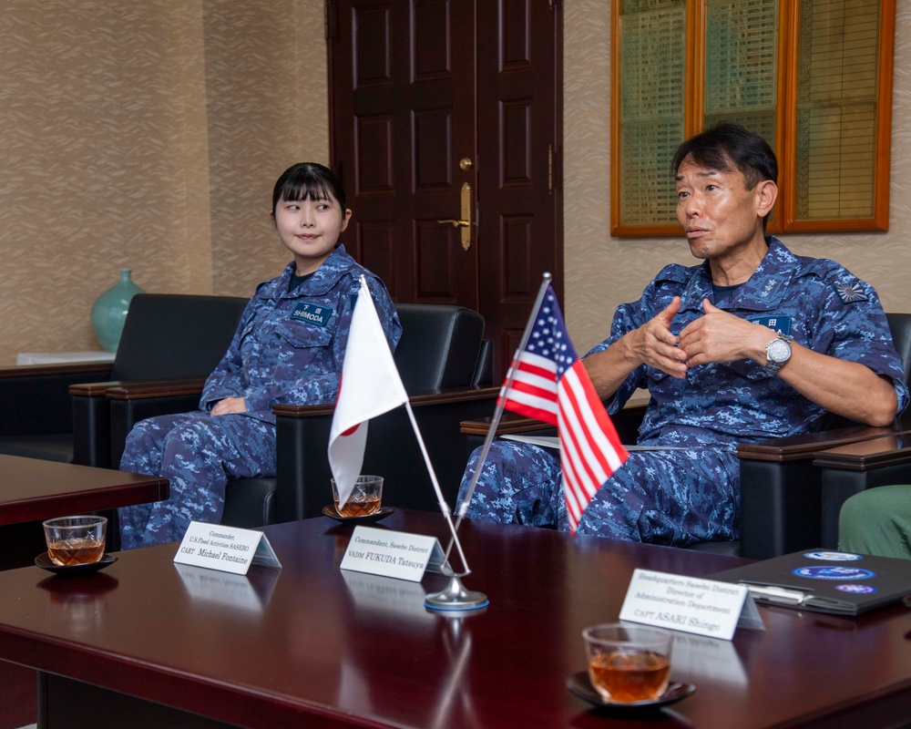 Two individuals in Japanese Maritime Self-Defense Force (JMSDF) digital camouflage uniforms are seated at a conference table during what appears to be an official bilateral meeting. Name placards on the table identify participants including VADM FUKUDA Tatsuya (Commandant, Sasebo District) and CAPT Michael Fontaine (Commander, U.S. Fleet Activities Sasebo). Both Japanese and American flags are displayed on the table, indicating a U.S.-Japan military liaison meeting.