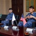 Two individuals in Japanese Maritime Self-Defense Force (JMSDF) digital camouflage uniforms are seated at a conference table during what appears to be an official bilateral meeting. Name placards on the table identify participants including VADM FUKUDA Tatsuya (Commandant, Sasebo District) and CAPT Michael Fontaine (Commander, U.S. Fleet Activities Sasebo). Both Japanese and American flags are displayed on the table, indicating a U.S.-Japan military liaison meeting.