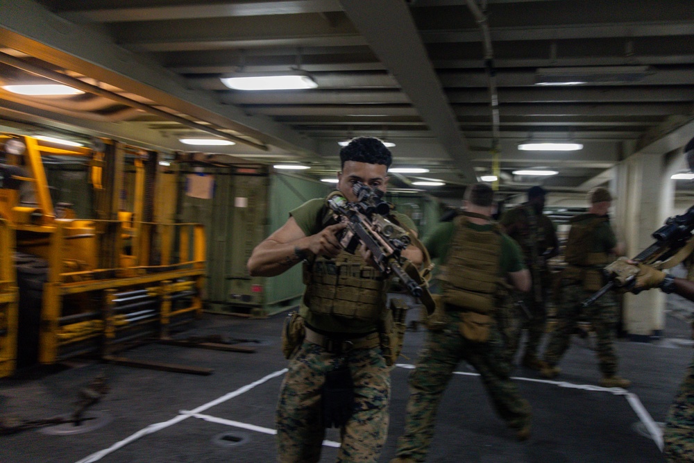 Military personnel in camouflage uniforms and tactical vests are conducting what appears to be a training exercise or drill inside a large enclosed space, likely the vehicle deck or hangar bay of a naval vessel. The lead figure is aiming a rifle directly toward the camera while moving forward with other personnel following behind. The environment features industrial equipment, overhead lighting, and painted deck markings consistent with a ship's interior.