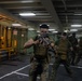 Military personnel in camouflage uniforms and tactical vests are conducting what appears to be a training exercise or drill inside a large enclosed space, likely the vehicle deck or hangar bay of a naval vessel. The lead figure is aiming a rifle directly toward the camera while moving forward with other personnel following behind. The environment features industrial equipment, overhead lighting, and painted deck markings consistent with a ship's interior.