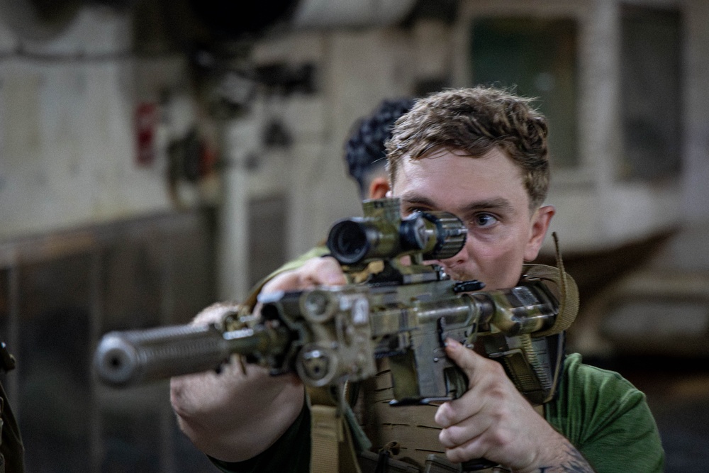 A young male in tactical gear is aiming a camouflage-patterned rifle equipped with a scope and suppressor directly toward the camera. He is wearing a plate carrier vest over a green shirt and appears to be in an indoor industrial or military environment. A second individual is partially visible in the background.