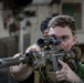 A young male in tactical gear is aiming a camouflage-patterned rifle equipped with a scope and suppressor directly toward the camera. He is wearing a plate carrier vest over a green shirt and appears to be in an indoor industrial or military environment. A second individual is partially visible in the background.