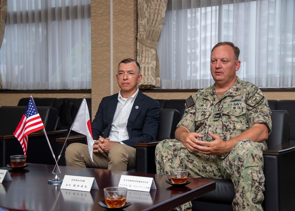 Two individuals are seated at a conference table during what appears to be a formal bilateral meeting between U.S. and Japanese officials. A U.S. Navy officer in camouflage uniform with the name tape 'FONTAINE' is speaking, accompanied by a civilian in a dark blazer. Small American and Japanese flags are displayed on the table alongside name placards with Japanese and English text.