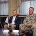 Two individuals are seated at a conference table during what appears to be a formal bilateral meeting between U.S. and Japanese officials. A U.S. Navy officer in camouflage uniform with the name tape 'FONTAINE' is speaking, accompanied by a civilian in a dark blazer. Small American and Japanese flags are displayed on the table alongside name placards with Japanese and English text.