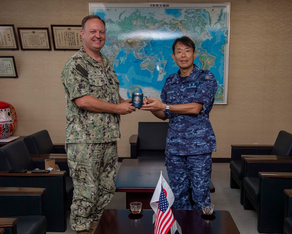 Two military officers, one in U.S. Navy digital camouflage (name tape: FONTAINE) and one in Japan Air Self-Defense Force blue digital camouflage (name tape: FUKUDA), are jointly holding a commemorative gift item during a formal bilateral meeting. The setting appears to be an official office in Japan, decorated with a world map in Japanese, framed certificates, and a traditional Daruma doll. Small Japanese and American flags on the table indicate a bilateral diplomatic or military exchange event.