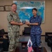 Two military officers, one in U.S. Navy digital camouflage (name tape: FONTAINE) and one in Japan Air Self-Defense Force blue digital camouflage (name tape: FUKUDA), are jointly holding a commemorative gift item during a formal bilateral meeting. The setting appears to be an official office in Japan, decorated with a world map in Japanese, framed certificates, and a traditional Daruma doll. Small Japanese and American flags on the table indicate a bilateral diplomatic or military exchange event.
