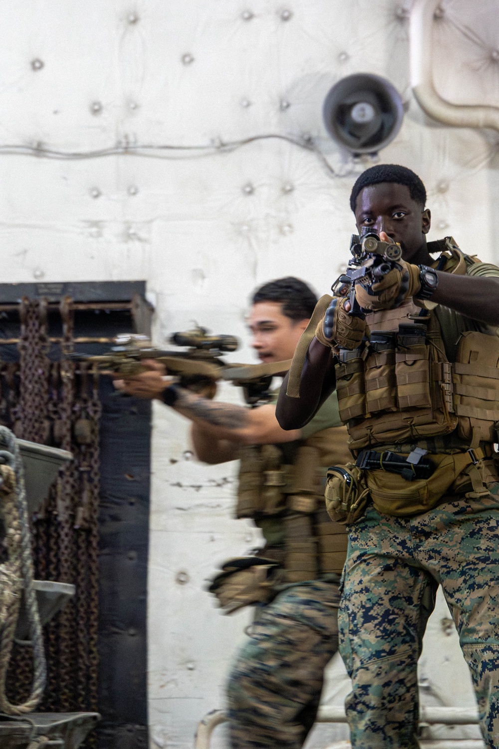Two military personnel in full tactical gear are conducting what appears to be a training exercise aboard a naval vessel or military ship interior. The foreground subject is aiming a rifle with an optic directly toward the camera, while a second operator moves in the background. Both are equipped with MOLLE plate carriers, camouflage uniforms consistent with USMC MARPAT pattern, and tactical accessories.