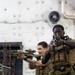 Two military personnel in full tactical gear are conducting what appears to be a training exercise aboard a naval vessel or military ship interior. The foreground subject is aiming a rifle with an optic directly toward the camera, while a second operator moves in the background. Both are equipped with MOLLE plate carriers, camouflage uniforms consistent with USMC MARPAT pattern, and tactical accessories.