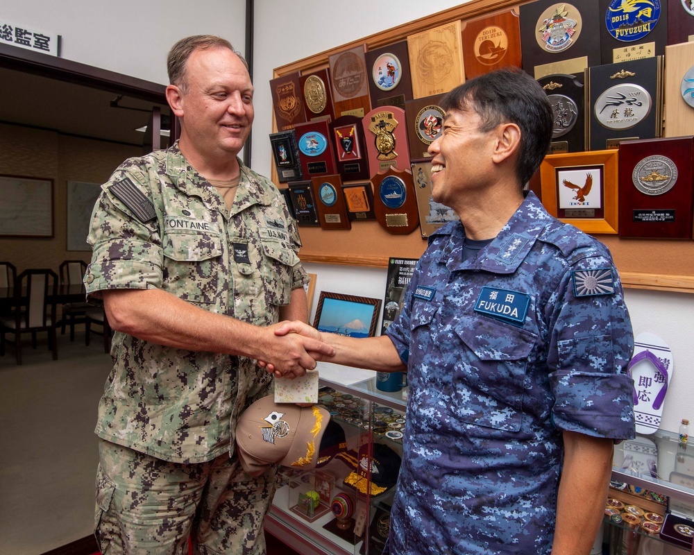 Two military officers are shaking hands in what appears to be an official meeting room. The officer on the left wears a U.S. Navy digital camouflage uniform with the name tag 'FONTAINE', while the officer on the right wears a Japan Maritime Self-Defense Force (JMSDF) blue digital camouflage uniform with the name tag 'FUKUDA'. The background displays numerous military plaques, unit crests, and commemorative items on the wall.