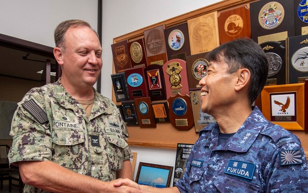 Two military officers are shaking hands in what appears to be an official meeting room. The officer on the left wears a U.S. Navy digital camouflage uniform with the name tag 'FONTAINE', while the officer on the right wears a Japan Maritime Self-Defense Force (JMSDF) blue digital camouflage uniform with the name tag 'FUKUDA'. The background displays numerous military plaques, unit crests, and commemorative items on the wall.