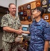 Two military officers are shaking hands in what appears to be an official meeting room. The officer on the left wears a U.S. Navy digital camouflage uniform with the name tag 'FONTAINE', while the officer on the right wears a Japan Maritime Self-Defense Force (JMSDF) blue digital camouflage uniform with the name tag 'FUKUDA'. The background displays numerous military plaques, unit crests, and commemorative items on the wall.